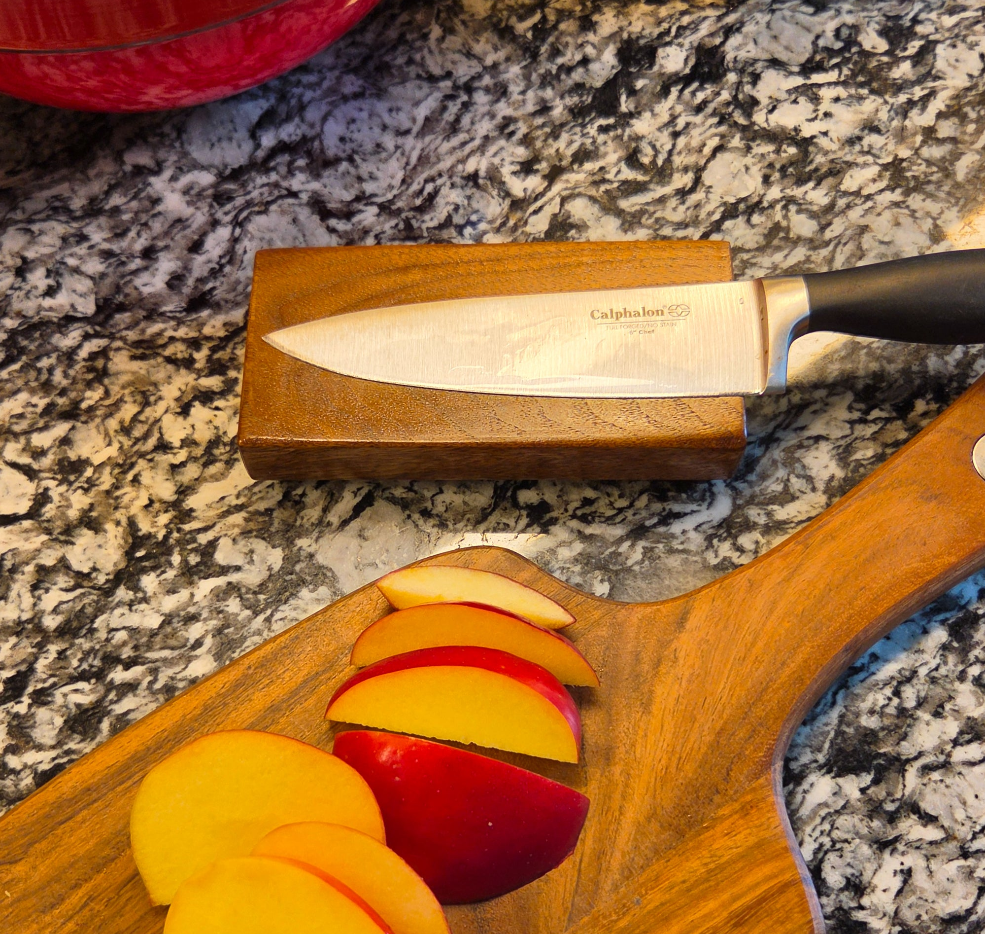 A Calphalon chef's knife with a black handle rests on a small wooden block, set on a speckled granite countertop. In the foreground, a wooden cutting board holds sliced red and yellow fruit.