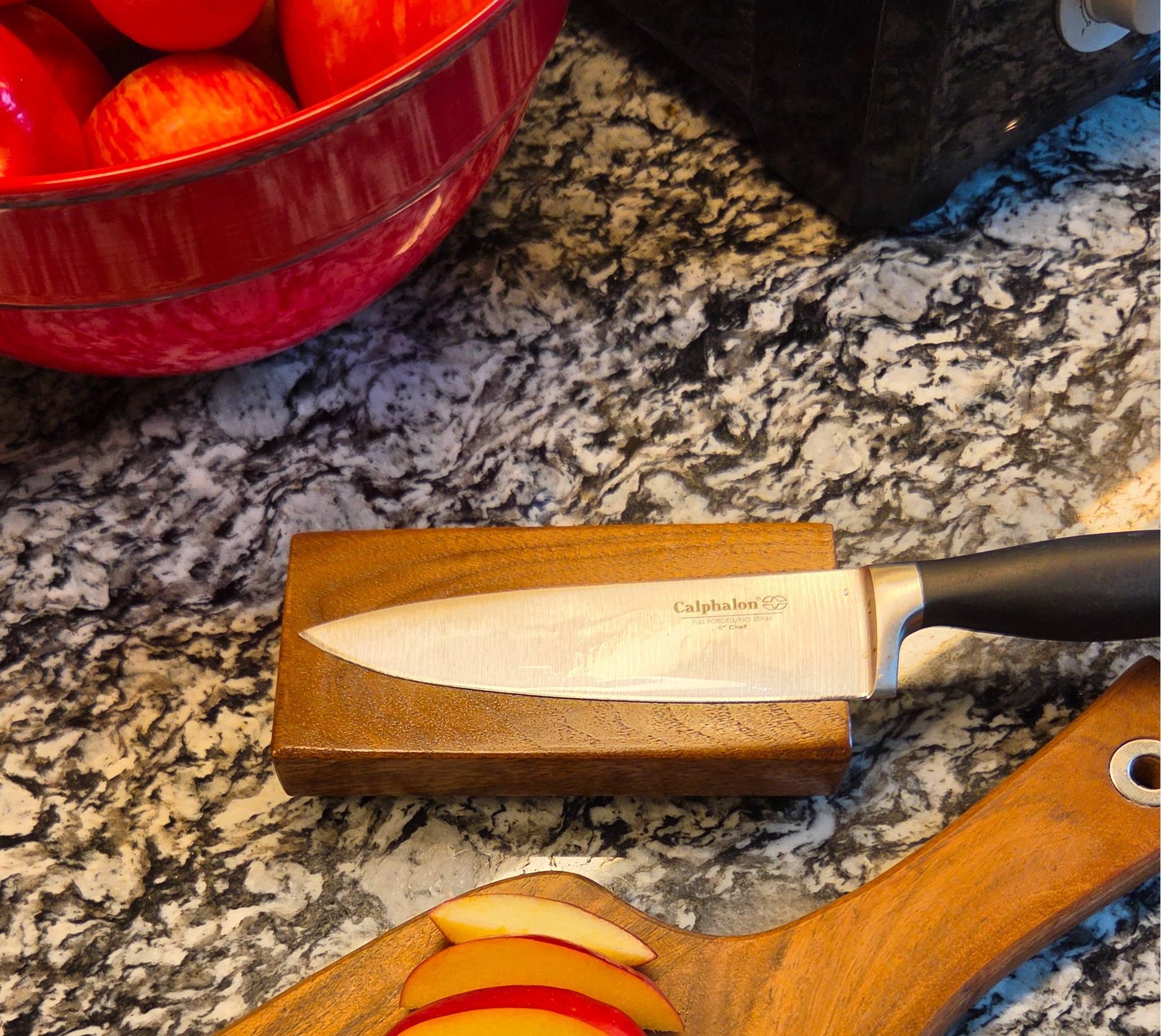 A Calphalon chef's knife with a black handle rests on a wooden block over a granite countertop, with sliced apples on a wooden cutting board and a red bowl of apples in the background.