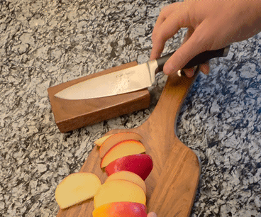 A hand holding a chef's knife over a wooden cutting board with sliced apples, next to a wooden block on a speckled granite countertop.